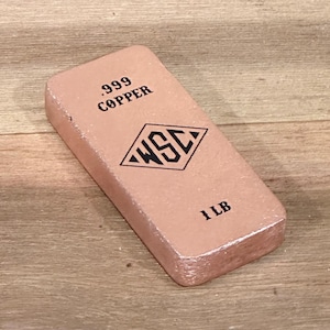 May include: A copper bar with the text ".999 COPPER" and the letters "WSC" inside a diamond shape. The bar weighs 1 LB. The bar is a light reddish-brown color and sits on a wooden surface.