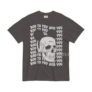 Może przedstawiać: Ciemnoszary t-shirt z kr&oacute;tkim rękawem z centralną grafiką jasnoszarej ludzkiej czaszki. Wz&oacute;r otoczony jest powtarzającym się białym napisem "BOO TO YOU AND YOU", ułożonym w falisty, prostokątny wz&oacute;r na wysokości klatki piersiowej.