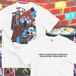 Może przedstawiać: Biały t-shirt z grafiką na plecach przedstawiającą klauna, namiot cyrkowy, balony i znak z napisem DOWNTOWN CLOWN TOWN. Z przodu koszulki znajduje się czarny napis I WENT TO DOWNTOWN CLOWNTOWN AND EVERYONE THERE KNEW YOU.