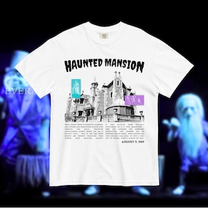 Może przedstawiać: Biały t-shirt z krótkim rękawem z czarno-białą grafiką przedstawiającą posiadłość oraz napisem HAUNTED MANSION u góry. Wzór zawiera małe niebieskie i fioletowe prostokątne akcenty oraz nadrukowany tekst z datą 9 sierpnia 1969 roku.