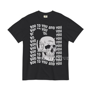 Może przedstawiać: Czarny t-shirt z kr&oacute;tkim rękawem z centralną białą grafiką przedstawiającą ludzką czaszkę. Tło grafiki składa się z powtarzającego się białego napisu "BOO TO YOU AND YOU" ułożonego w falisty, zniekształcony wz&oacute;r wok&oacute;ł czaszki.