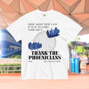 Może przedstawiać: Biały t-shirt z krótkim rękawem z czarną grafiką przedstawiającą sferę geodezyjną i dwa niebieskie pojazdy z atrakcji. Tekst głosi: THINK ABOUT HOW EASY IT WAS TO LEARN YOUR ABC'S... THANK THE PHOENICIANS 1982 THEY INVENTED THEM!