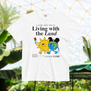 Może przedstawiać: Biały t-shirt z krótkim rękawem z grafiką przedstawiającą żółte słońce i niebieską ziemię trzymające się za ręce. Tekst głosi: I would rather truly be Living with the Land, Welcome to the Future Community of Tomorrow, September 1982.