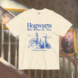 May include: A white t-shirt with a blue graphic of Hogwarts castle and the text "Hogwarts Will Always Be There To Welcome You Home".