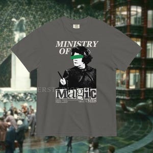 May include: A gray t-shirt with the words "MINISTRY OF MAGIC" above a black and white graphic of a person with green eye coverings holding a wand. The word "Magic" is below the graphic.