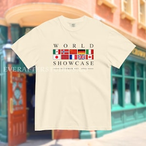 Może przedstawiać: Kremowa koszulka z napisem "WORLD SHOWCASE" nad rzędem flag międzynarodowych. Data "OCTOBER 1ST, 1982" jest wydrukowana poniżej. Koszulka ma krótkie rękawy i okrągły dekolt.