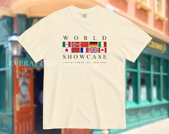 May include: Cream-colored t-shirt featuring the text "WORLD SHOWCASE" above a row of international flags. The date "OCTOBER 1ST, 1982" is printed below the text. The shirt has short sleeves and a crew neck.