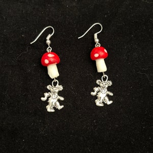 Könnte beinhalten: Ein Paar silberne Ohrringe mit einem roten und weißen Pilz-Anhänger und einem silbernen Bären-Anhänger, der darunter hängt.