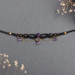 Puede incluir: Un collar gargantilla de macramé negro con piedras de amatista y detalles dorados. El collar presenta una cuenta central de amatista, gotas de amatista colgantes y detalles de cadena dorada, creando un estilo bohemio elegante.