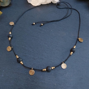 May include: A black cord necklace featuring alternating round black lava beads and small metallic gold-toned beads. The necklace is accented with several circular gold-toned charms featuring concentric ring patterns and has an adjustable cord closure.