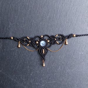 May include: A black macrame choker necklace featuring intricate knotted cord patterns, a central round light blue cabochon stone, small metallic gold-toned beads, and delicate gold-toned chain accents with three small dangling metallic charms.