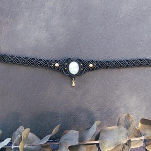 Könnte beinhalten: Ein schwarzes Makramee-Choker-Halsband mit einem ovalen, hellfarbigen Mittelstein. Die Halskette hat goldfarbene Akzente und einen kleinen, baumelnden Anhänger. Das aufwendige Webdesign verleiht diesem handgefertigten Schmuck einen Bohemian-Touch.
