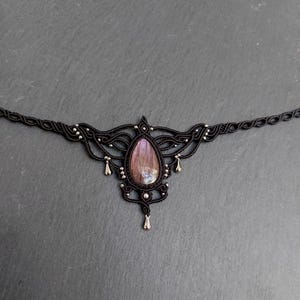 May include: A black macramé choker necklace with a central, teardrop-shaped, iridescent purple gemstone. The necklace features detailed knotwork, small silver beads, and dangling accents, offering a delicate, bohemian style.