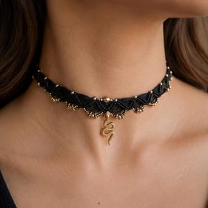 May include: A black macrame choker necklace featuring a central gold-toned snake pendant. The band is intricately woven with black cord and accented with small, evenly spaced gold-toned beads along the lower edge, creating a textured design.
