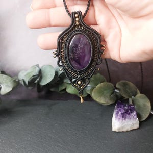 Op de afbeelding: Een macramé hanger ketting met een grote, ovaalvormige paarse amethist edelsteen gezet in ingewikkeld zwart koordvlechtwerk. Het ontwerp bevat kleine goudkleurige metalen kralen verwerkt in het weefsel en een enkele goudkleurige druppelbedel.