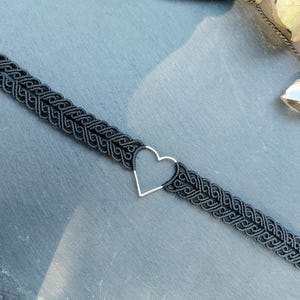 Puede incluir: Collar gargantilla de macramé negro con un colgante de corazón plateado. El collar presenta un diseño tejido y se muestra sobre una superficie gris. El colgante de corazón añade un toque de elegancia al accesorio hecho a mano.