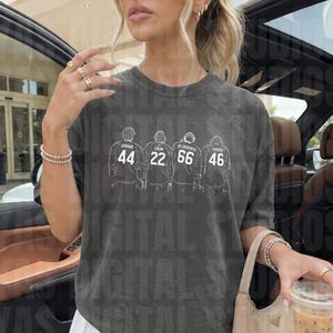 May include: A charcoal grey short-sleeve t-shirt featuring a white line-art graphic of four figures in jerseys with the names Graham 44, Logan 22, De Laurny 66, and Tucker 46 printed above their respective numbers on the back of the shirts.