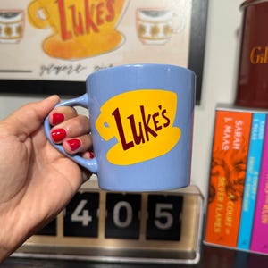 Peut inclure: Une tasse en céramique bleu clair ornée d'un motif jaune représentant une tasse à café avec l'inscription "Luke's" en lettres rouge foncé. La tasse possède une anse incurvée sur le côté gauche et une finition extérieure lisse et unie.
