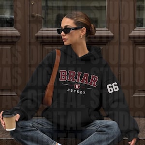 May include: A black hooded sweatshirt featuring the text BRIAR U HOCKEY in maroon and white lettering on the chest, with the number 66 in white on the left sleeve. The garment is paired with dark grey denim jeans, white trainers, and a brown shoulder bag.