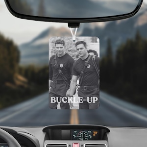 May include: A rectangular car air freshener hanging from a rearview mirror, featuring a black and white photograph of two individuals in uniform with the text BUCKLE-UP printed in white capital letters at the bottom of the image.