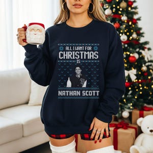 Op de afbeelding: Marineblauwe kersttrui met de tekst "ALL I WANT FOR CHRISTMAS IS NATHAN SCOTT" in wit en lichtblauw. De trui heeft een portret van Nathan Scott en sneeuwvlokontwerpen.