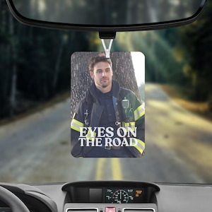 May include: A rectangular car air freshener hanging from a rearview mirror, featuring a photograph of a firefighter in a dark uniform with yellow reflective stripes, with the white text EYES ON THE ROAD printed across the lower portion of the image.
