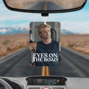 May include: A rectangular car air freshener hanging from a rearview mirror, featuring a photographic print of a blonde person in a dark sleeveless shirt with the white text EYES ON THE ROAD printed across the chest area of the garment.