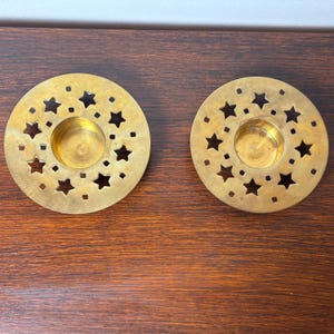 May include: Two circular brass candle holders featuring a flat, wide rim with a die-cut pattern of various sized stars and small squares. Each holder has a central, recessed circular cup designed to securely hold a single tea light candle.