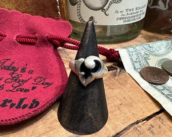 Può includere: Un anello d'argento con un design unico e vorticoso è esposto su un cono di legno scuro. È visibile un sacchetto rosso con coulisse con la scritta "Today is a Good Day to Love". Una bottiglia di rum è sullo sfondo.