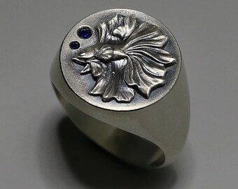 May include: A silver ring featuring an engraved design of a betta fish, with blue gemstones set on the top.