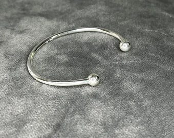 Op de afbeelding: Een zilveren armband met twee ronde uiteinden. De armband ligt op een grijs stoffen oppervlak.