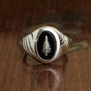 Puede incluir: Un anillo de plata con una piedra ovalada negra que presenta un diseño de árbol plateado.