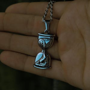 Könnte beinhalten: Eine silberfarbene Halskette mit Anhänger, die ein Sanduhr-Design mit einem Totenkopf am unteren Ende aufweist. Die Sanduhr hat eine detaillierte, gotische Ästhetik. Die Kette ist ebenfalls silberfarben und besteht aus kleinen, verbundenen Teilen. Die Halskette wird in einer Hand gehalten.