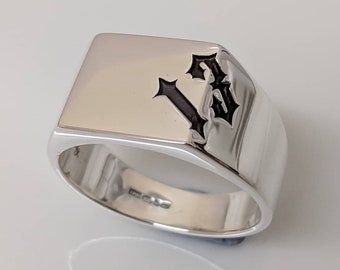 Può includere: Un anello chevalier in argento lucido con una faccia rettangolare. L'anello presenta un design nero a forma di lettera "B" in stile gotico. L'anello ha una fascia larga.