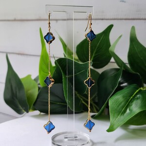 May include: A pair of long gold dangle earrings with three blue four-leaf clover charms on each earring.