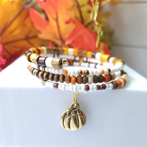 May include: A multi-strand wrap bracelet with a pumpkin charm. The bracelet features wooden beads in shades of brown, orange, white, and cream. The gold-colored charm is a detailed pumpkin. The bracelet is displayed on a white surface with fall foliage in the background.