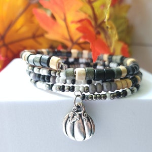 May include: A multi-strand wrap bracelet with a silver pumpkin charm. The bracelet features a mix of wooden beads in black, cream, and gray, along with smaller metallic beads. The background includes fall foliage.