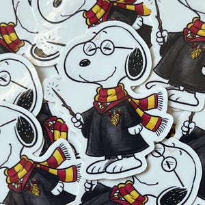 May include: A collection of white Snoopy stickers, each dressed in a black robe, a red and yellow striped scarf, and round glasses, holding a wand. The stickers feature the Gryffindor crest and are scattered across a surface.
