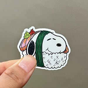 May include: A white Snoopy sticker designed as sushi, with a green seaweed wrap and rice. The sushi has salmon and wasabi toppings. The sticker is held by a hand against a gray background.