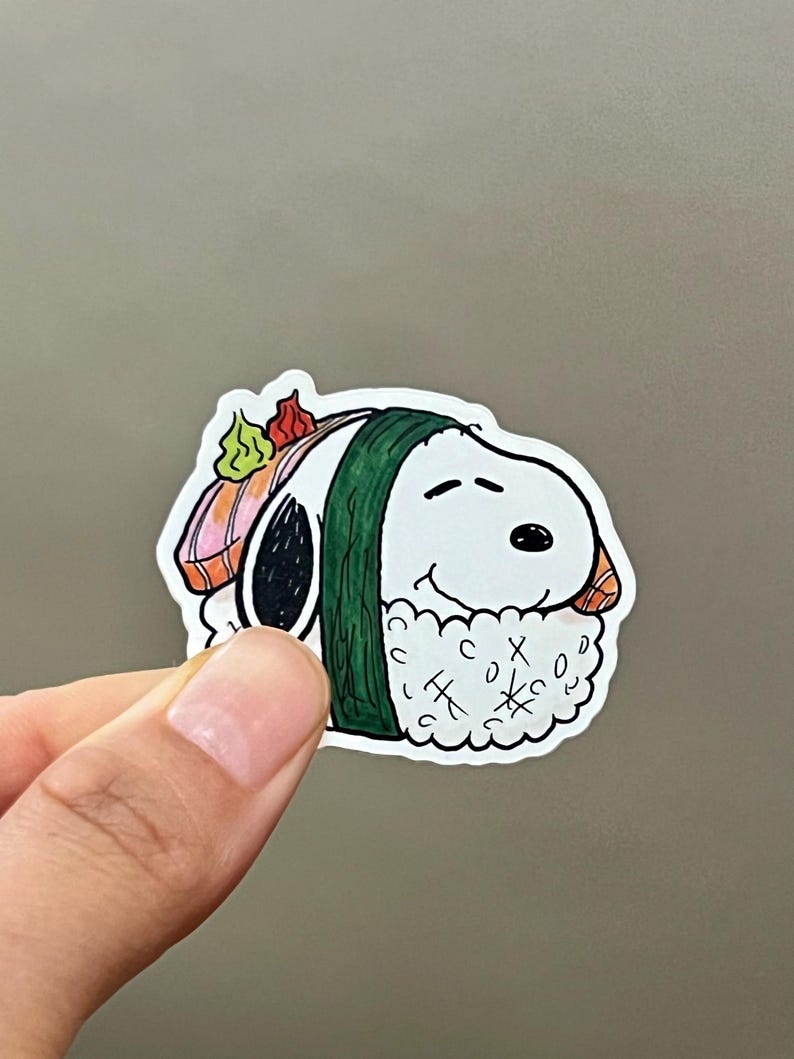 May include: A white Snoopy sticker designed as sushi, with a green seaweed wrap and rice. The sushi has salmon and wasabi toppings. The sticker is held by a hand against a gray background.