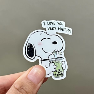 Pode incluir: Um adesivo branco de Snoopy com detalhes em preto, bebendo ch&aacute; de bolhas verde. O adesivo tem o texto "I LOVE YOU VERY MATCHA" em um bal&atilde;o de fala. Snoopy tem um rubor rosa nas bochechas.
