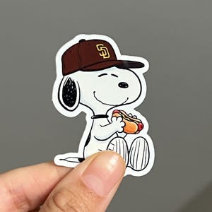 May include: A white cartoon character wearing a brown baseball cap with a gold "SD" logo, holding a hot dog. The character is smiling and has black ears and a black collar. The sticker is on a gray background.