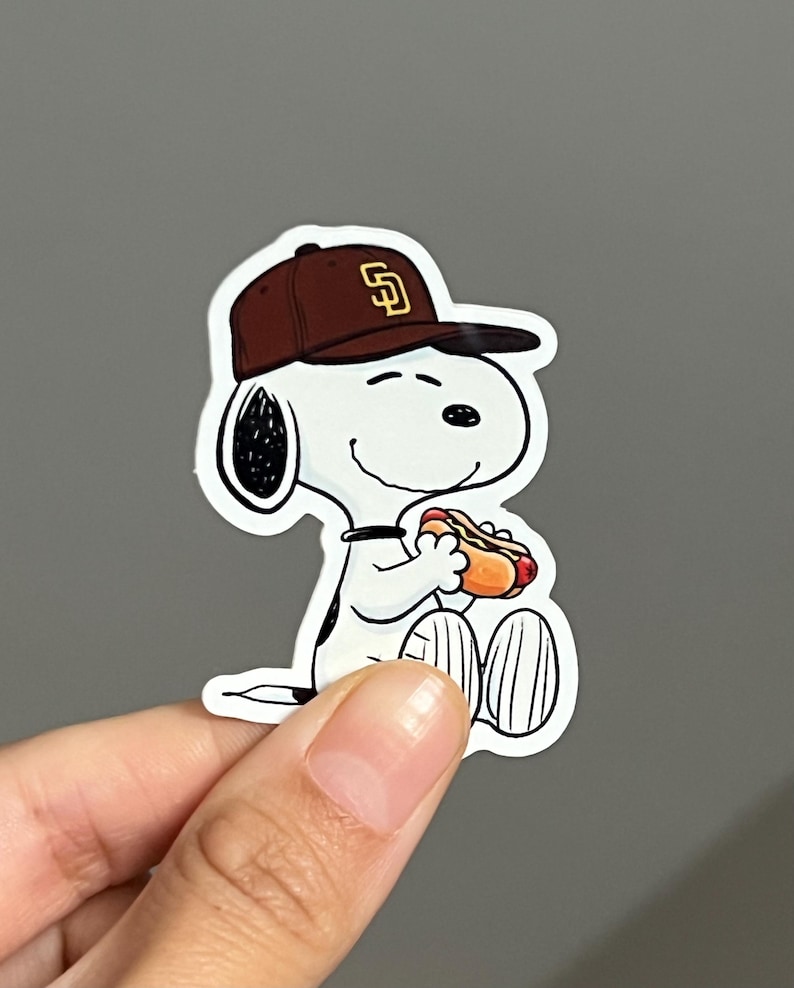 May include: A white cartoon character wearing a brown baseball cap with a gold "SD" logo, holding a hot dog. The character is smiling and has black ears and a black collar. The sticker is on a gray background.