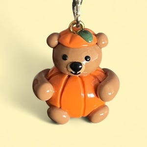 May include: A brown teddy bear pendant wearing an orange pumpkin costume. The bear has a black nose and eyes, and a small green leaf on its pumpkin hat. The pendant is attached to a silver chain.
