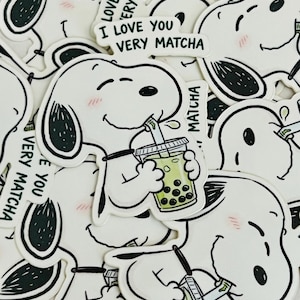 Pode incluir: Autocolantes brancos com um c&atilde;o de desenho animado com orelhas pretas e bochechas rosadas, bebendo ch&aacute; de bolhas. Os autocolantes t&ecirc;m o texto "I LOVE YOU VERY MATCHA" em verde.