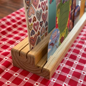 以下が含まれることがあります： スロット付きの木製カードホルダー。いくつかのグリーティングカードが表示されています。1枚のカードにはカラフルなハートが、他のカードには恐竜のイラストが描かれています。ホルダーは赤と白のチェック柄のテーブルクロスの上にあります。