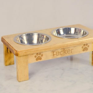 May include: A light-colored wooden pet feeder with two stainless steel bowls. The feeder has paw print cutouts and the name "Tucker" engraved on the front. The feeder is elevated on four legs.