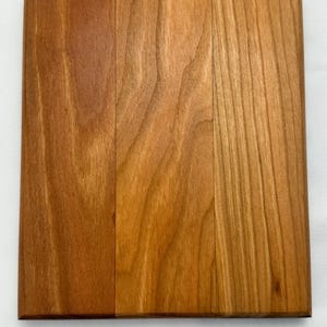 May include: A square wooden chopping board in a warm, reddish-brown colour. The wood grain is visible, and the board is divided into three vertical sections. The edges are cleanly cut, and the surface appears smooth.