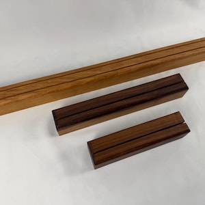 Puede incluir: Tres expositores de madera de diferentes tamaños. El más grande es de color marrón claro, mientras que los otros dos son de un marrón más oscuro. Cada soporte tiene una ranura en el centro.