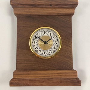 May include: A rectangular wooden clock with a gold-colored clock face. The clock face has black numerals and ornate detailing. A black metal ring is attached to the top for hanging. The wood has a rich, dark brown color.